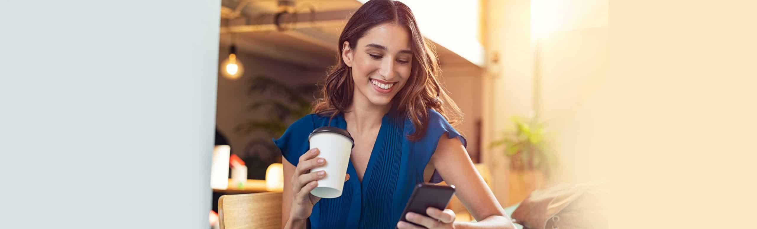 Woman holding a coffee cup smiling at her smartphone