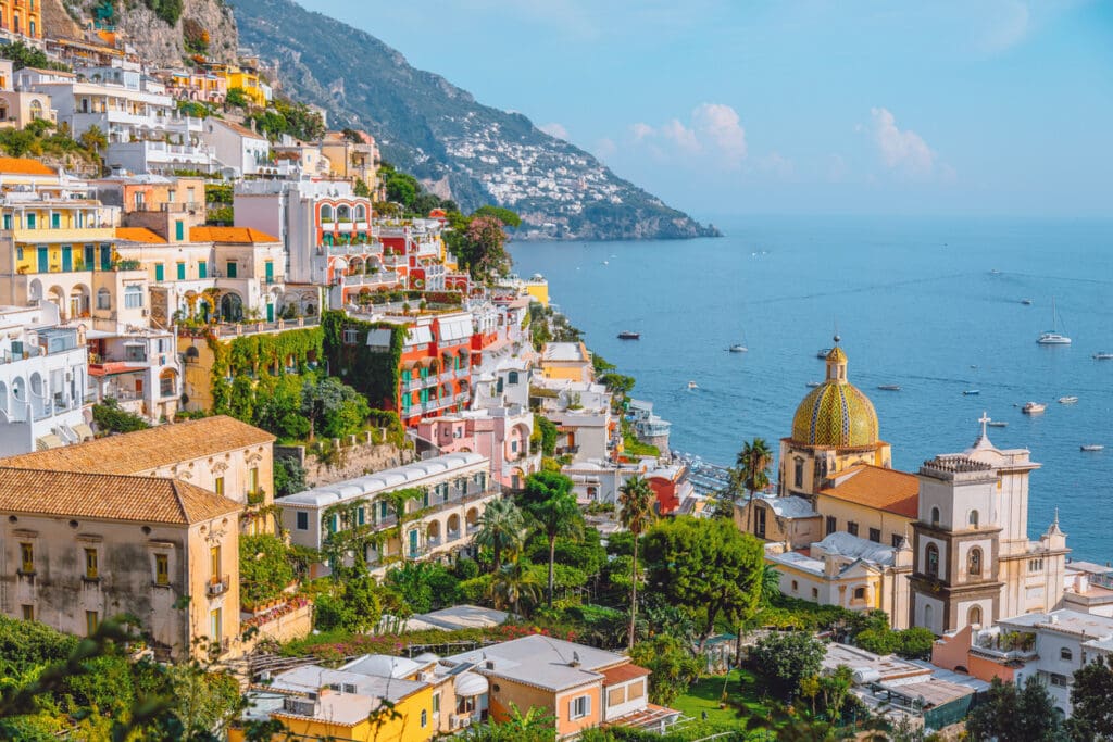 Detailed view of Positano’s iconic cliffside town with vibrant pastel houses, lush greenery, and the famous majolica-tiled dome of the Church of Santa Maria Assunta overlooking the Mediterranean Sea. Boats scatter across calm blue waters while terraces, palm trees, and sunlit buildings create a rich and layered composition of this world-renowned Italian coastal destination in Italia