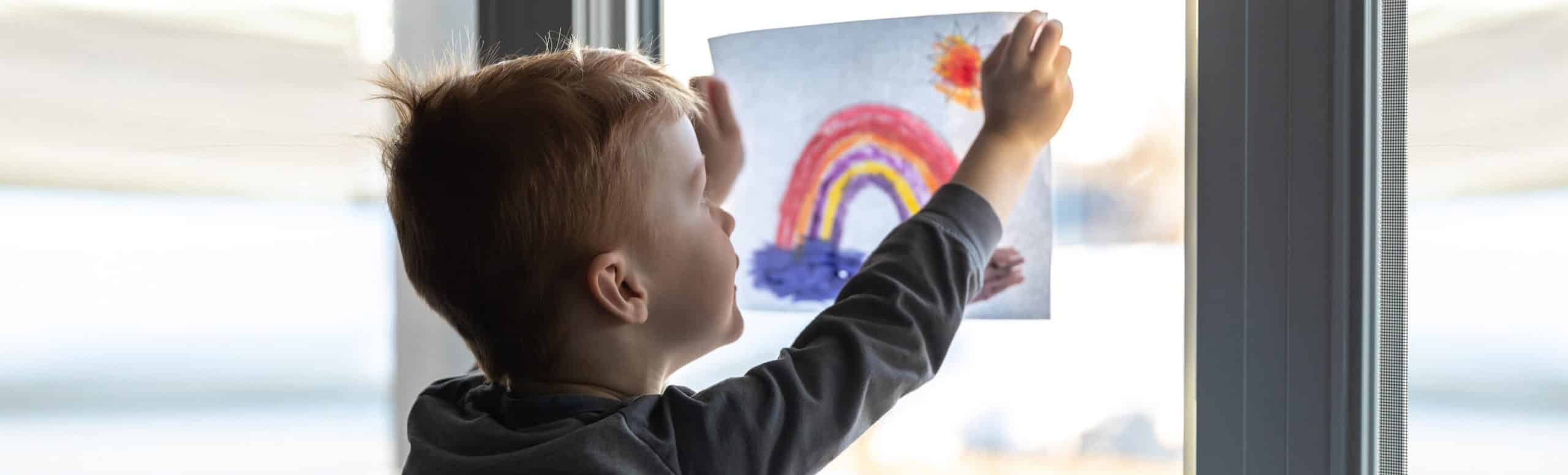 A kid sticking a drawing of a rainbow towards the window.