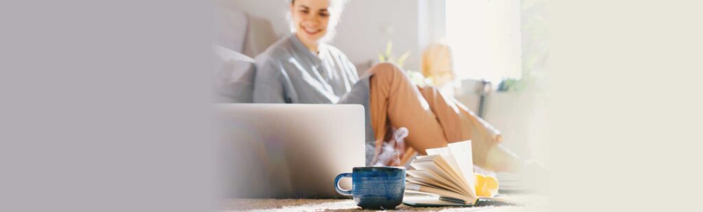 Person sitting on a carpet while working on her laptop with a mug and book in the foreground
