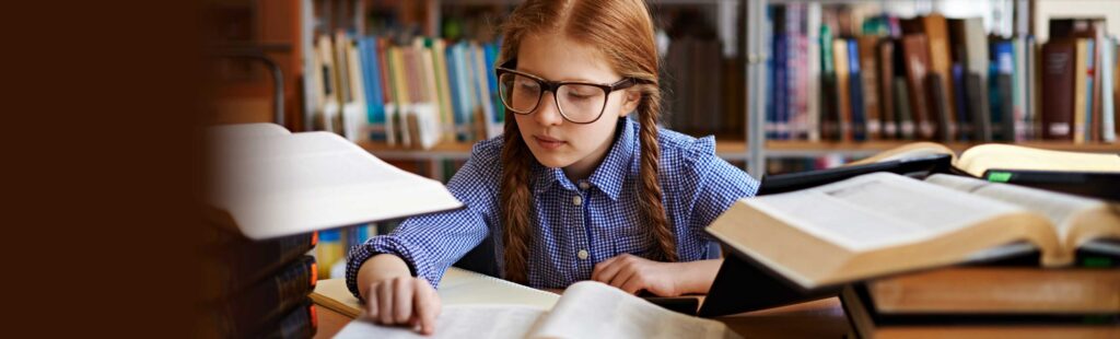 Young girl in braids and glasses reading a book while surrounded by many open books