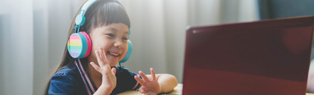 Child smiling with headphones online while waving to computer screen