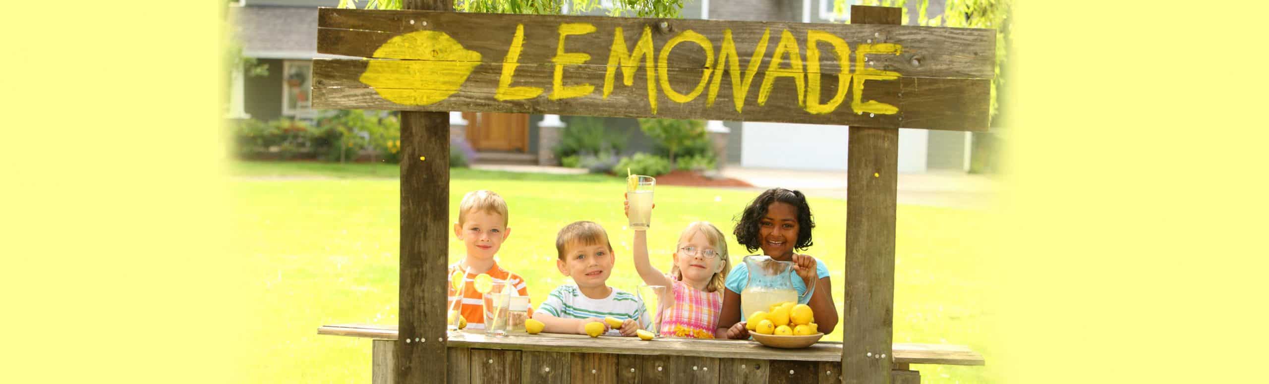 4 children behind a wooden lemonade stand with a sign that reads "LEMONADE"
