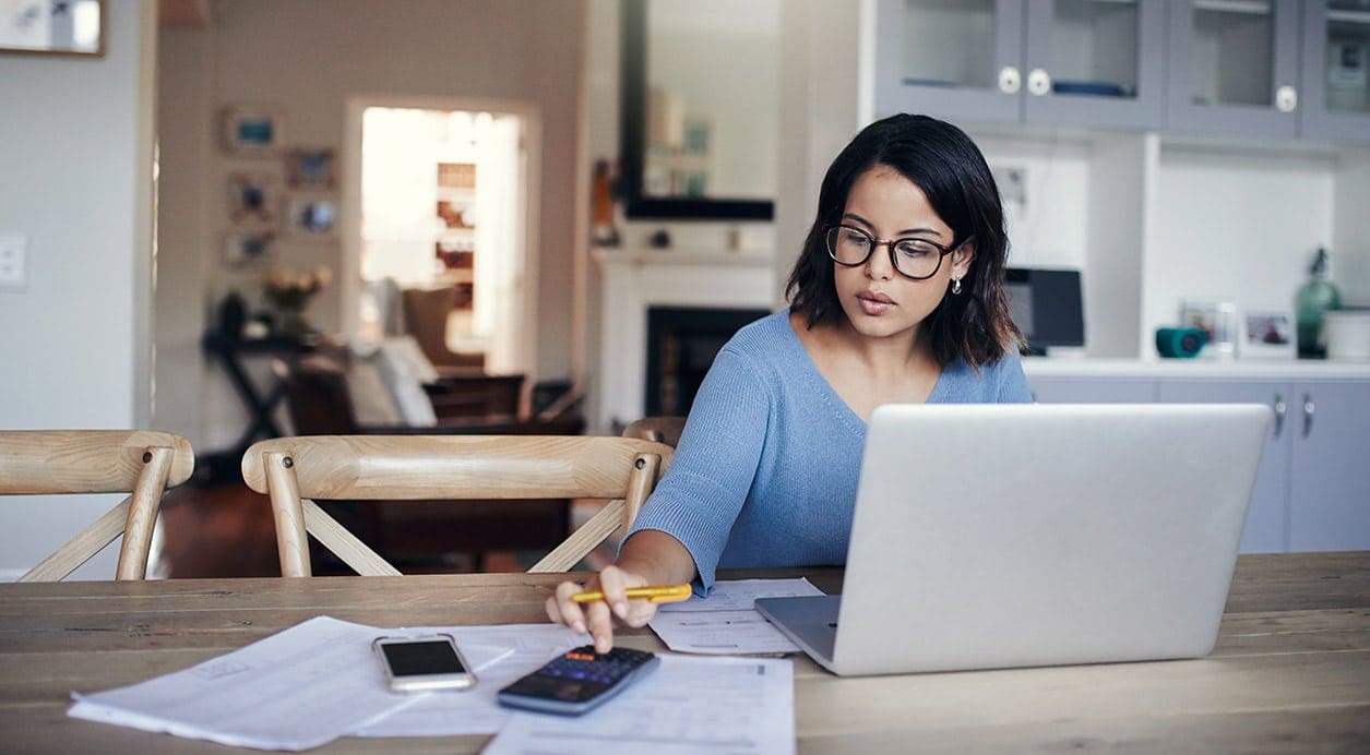 woman with glasses looking at phone on table while working on her laptop