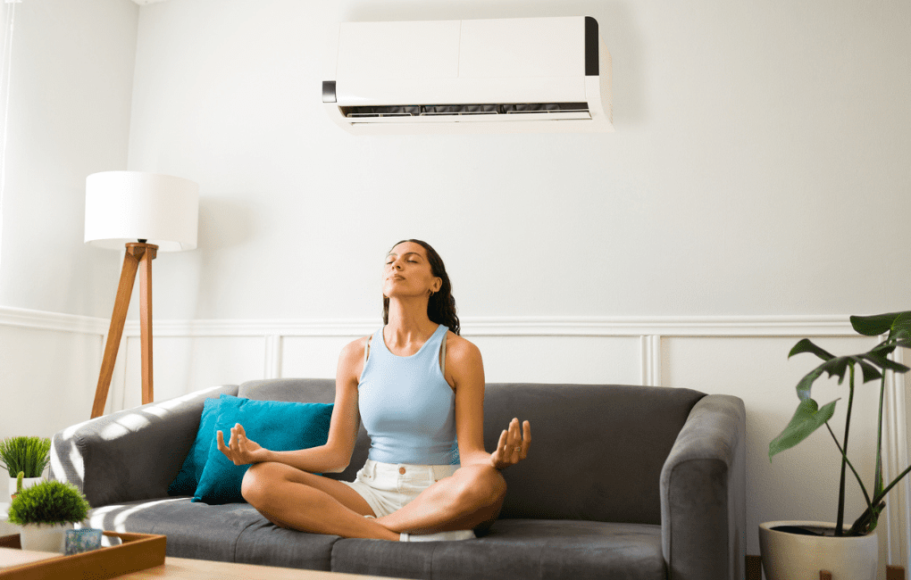 a woman sits in a zen pose on a couch under a ductless head pump in her living room