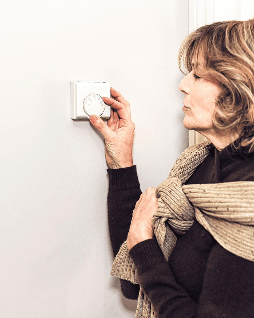 A woman with a sweater around her shoulders adjusts her home thermostat